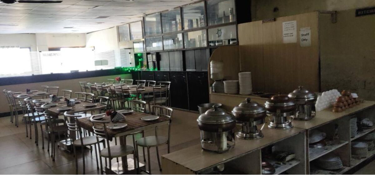 A cafeteria with tables and chairs, a buffet line with food containers, and a kitchen area in the background.