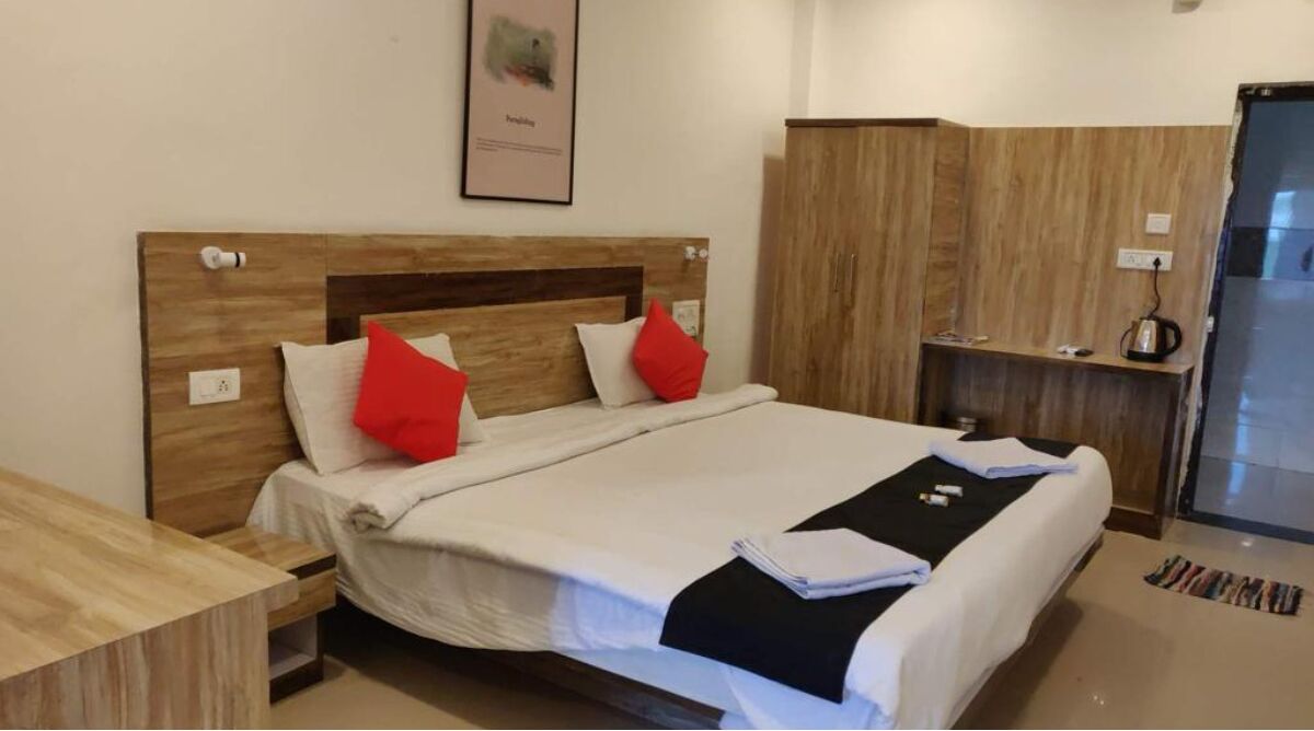 Una cama bien hecha con tablero de madera, almohadas rojas y una manta de color negro. A la derecha hay un armario de madera y una mesita con una tetera encima.