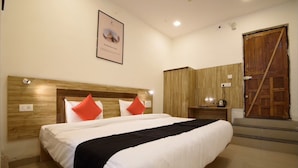 Een hotelkamer met een groot bed, een houten hoofdeinde, twee rode kussens, een zwarte plaid, een houten garderobe en een ingelijst schilderij aan de muur.