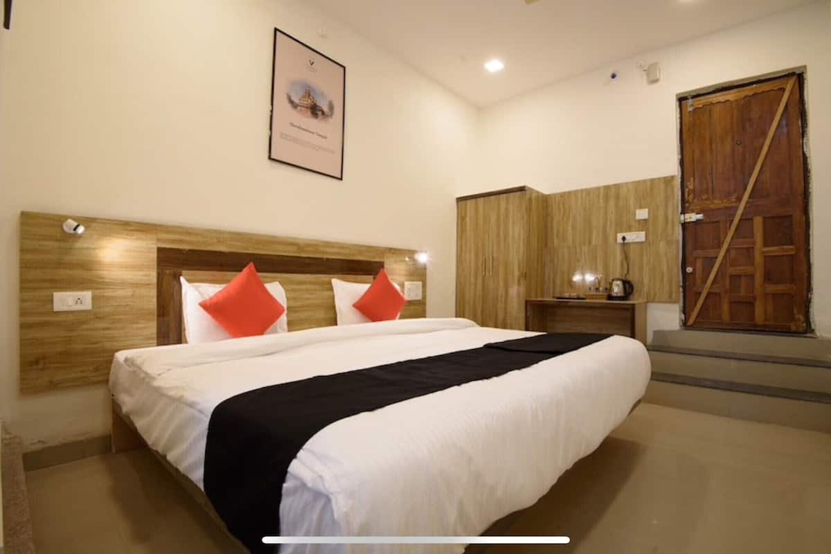 Une chambre d’hôtel avec un grand lit, une tête de lit en bois, deux oreillers rouges, un plaid noir, une armoire en bois et un tableau encadré au mur.