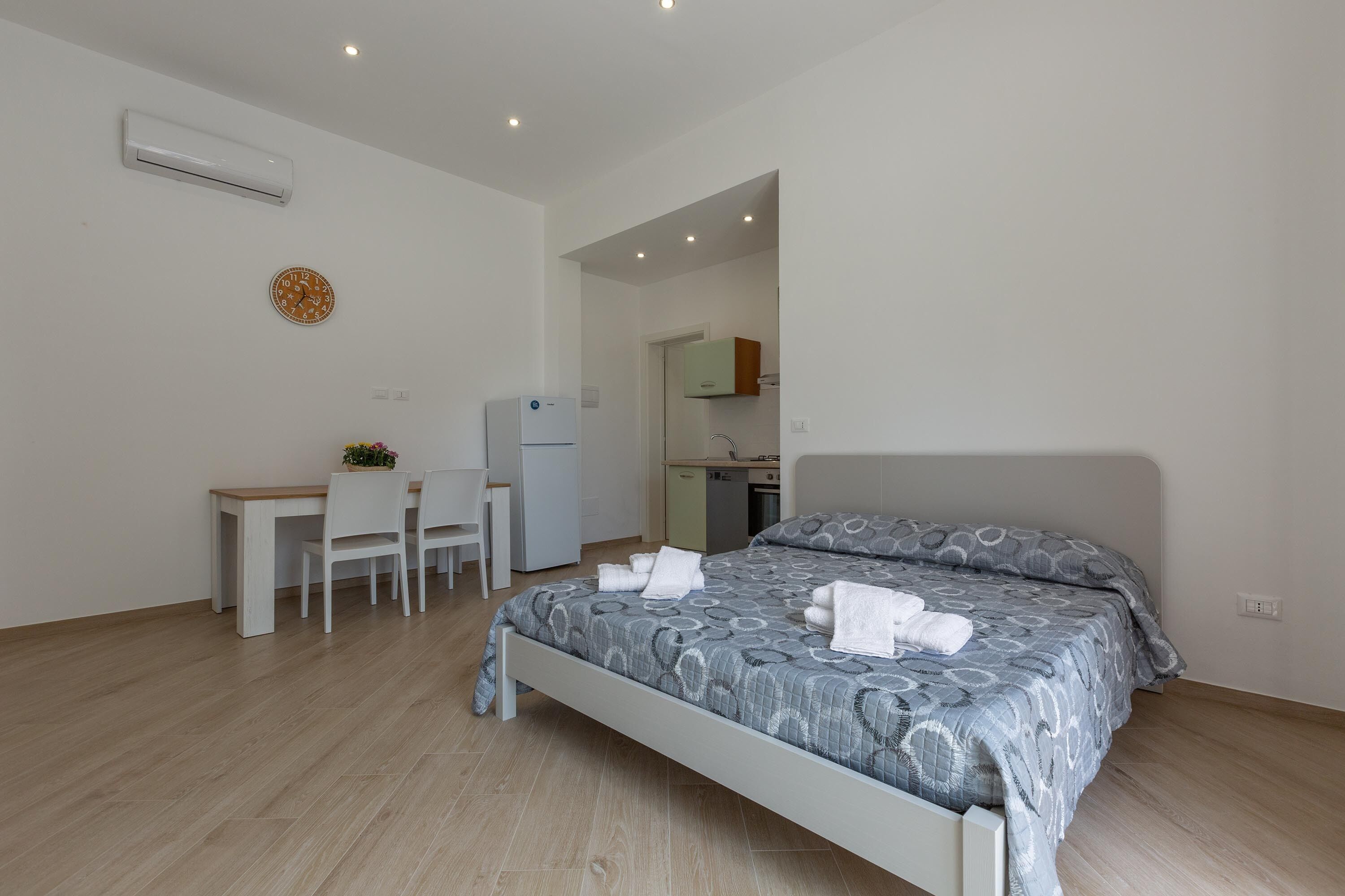 Een slaapkamer met een bed, een bureau en een stoel. Een keuken is zichtbaar door een open deur.