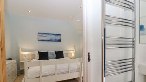 A bedroom with a bed, bedside tables, a nightstand, a wall-mounted painting, and a bathroom with a glass shower enclosure. - Mountfield Farm Cottage (Ashford)