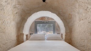 A bedroom with a stone archway, a large bed with white linens, and a window providing a view of the outdoors. - 3565 A Piedi Scalzi nel Trullo by Perle di Puglia (Latiano)