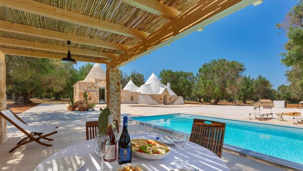 A dining table set with plates, cutlery, and bottles, overlooking a swimming pool and stone buildings. - 3565 A Piedi Scalzi nel Trullo by Perle di Puglia (Latiano)