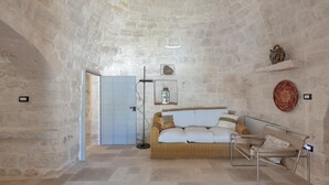 A room with a stone wall, a wicker sofa with cushions, a metal chair, a floor lamp, and a door. - 3565 A Piedi Scalzi nel Trullo by Perle di Puglia (Latiano)