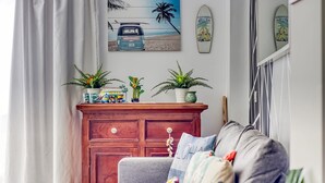 Una sala de estar con un sofá gris, un mueble de madera con plantas y un cuadro con motivos playeros en la pared.