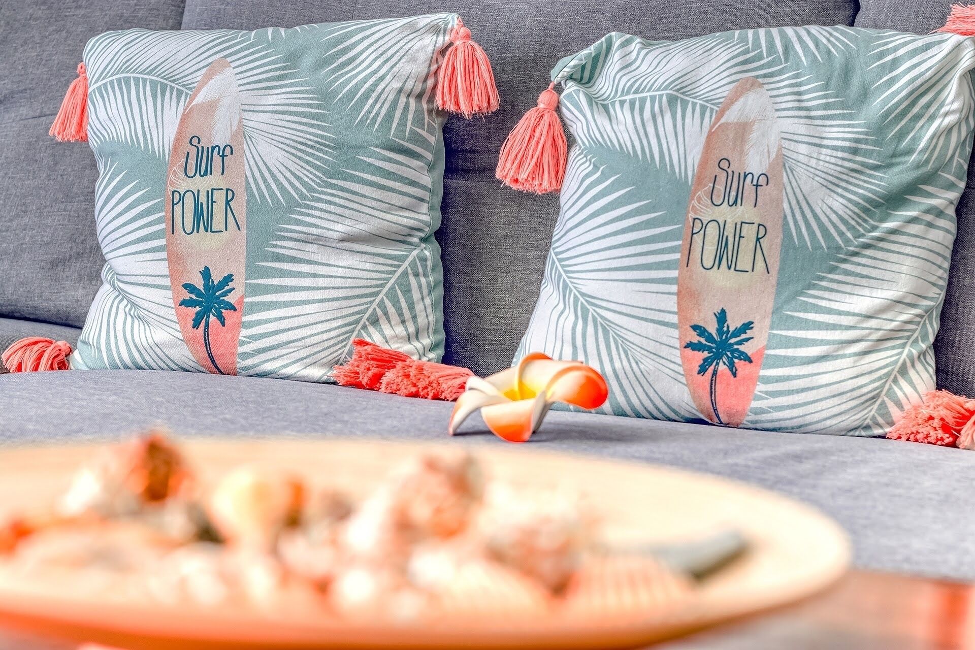 Duas almofadas com o texto 'Surf Power' e estampas de folhas de palmeira em um sofá cinza.
