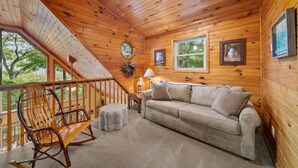 A wooden cabin interior with a rocking chair, a sofa, and a staircase leading to a balcony.