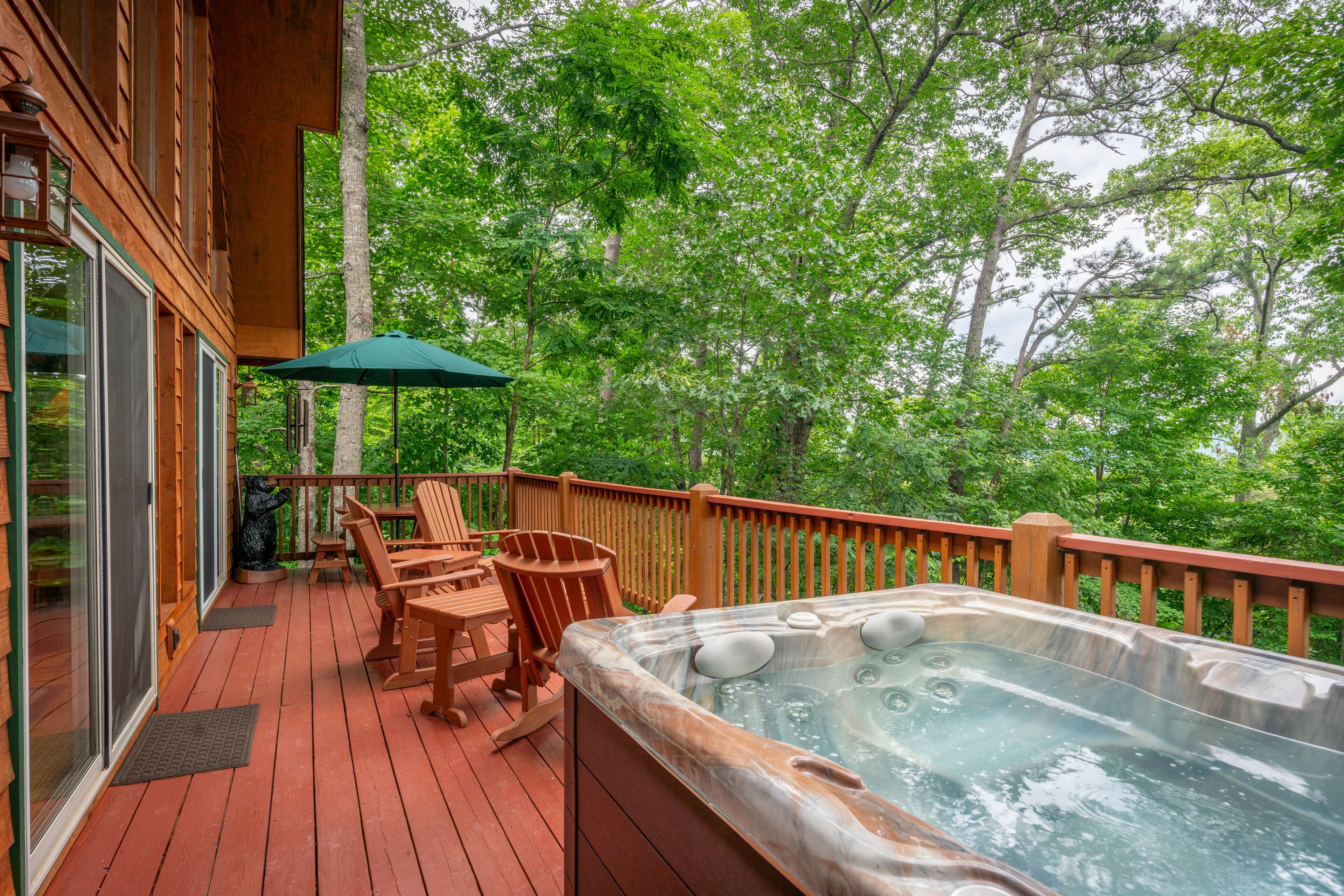 Een houten terras met een jacuzzi, buitenmeubels en uitzicht op de omliggende bomen.