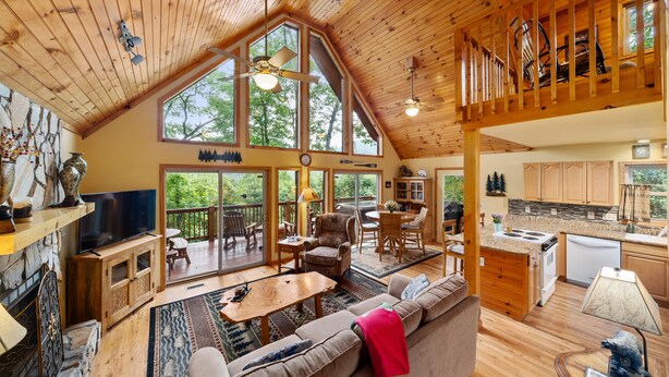 A spacious living area with a high ceiling, wooden beams, a large window, a TV, a fireplace, and a cozy seating area.