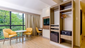 Une chambre d’hôtel dotée d’une grande fenêtre, d’un bureau avec des chaises, d’une télévision, d’un mini-réfrigérateur et d’une armoire où sont suspendus des vêtements.