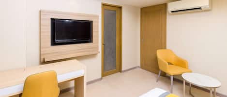 Quarto de hotel com televisão na parede, uma escrivaninha, duas cadeiras, uma mesinha e aparelho de ar condicionado.