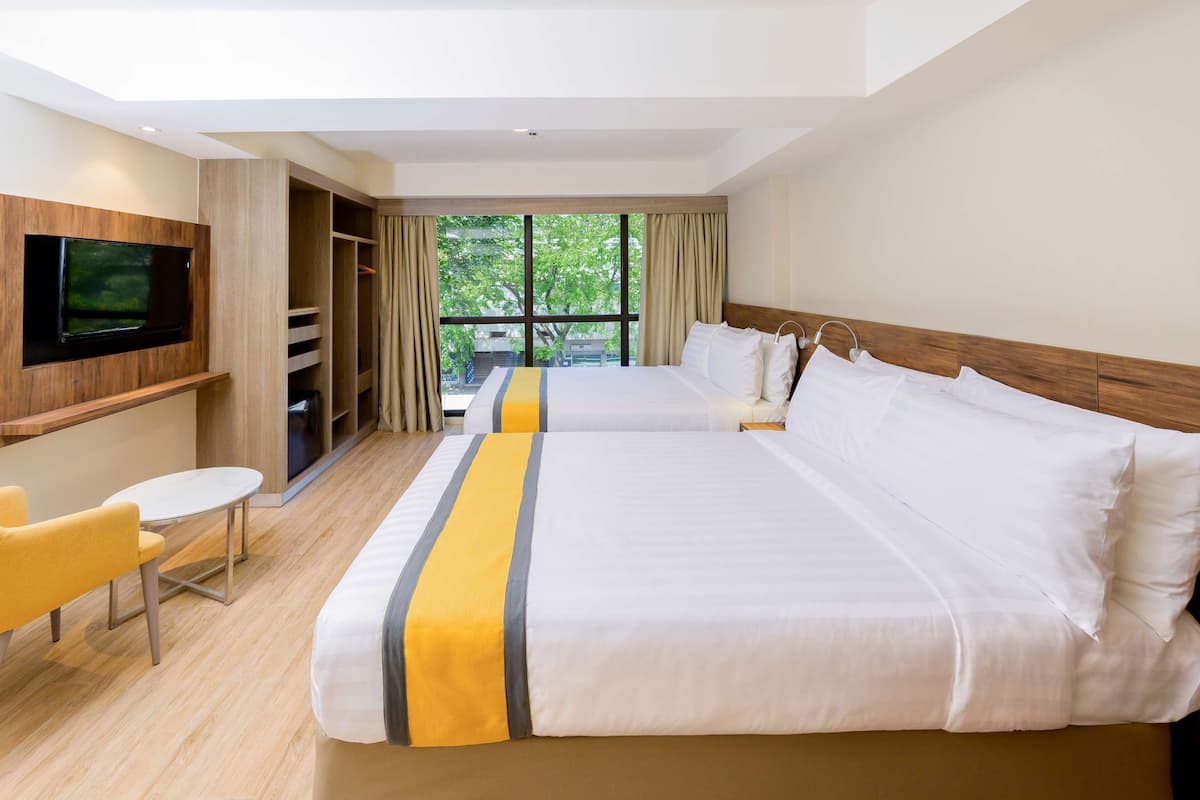 Ein Hotelzimmer mit zwei Betten, einem an der Wand befestigten Fernseher, einem kleinen Tisch und Stuhl sowie Blick auf Grünfläche.