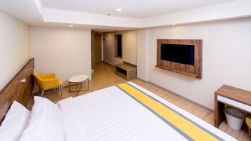 A hotel room with a bed, a TV mounted on the wall, a small seating area with a chair and table, and a trash bin.
