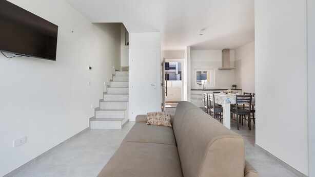 A modern living room with a beige sofa, a flat-screen TV mounted on the wall, and a staircase leading to a dining area with a table and chairs. - 3399 Bina House by Barbarhouse (Carovigno)