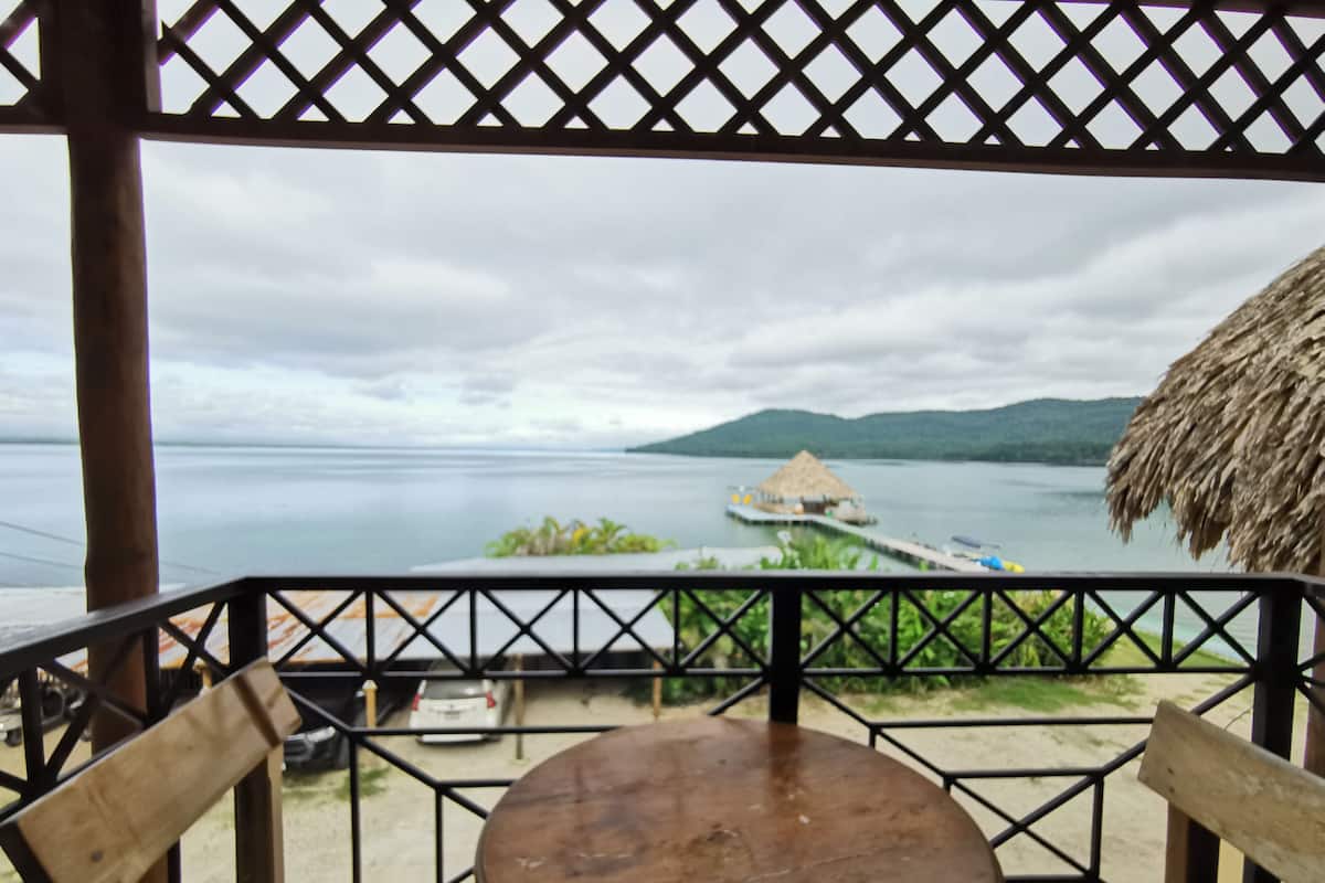 Una terraza de madera con una mesa y sillas, con vista a una masa de agua tranquila que incluye una estructura con techo de paja y un muelle que se adentra en el agua.