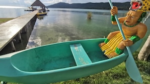 A green canoe with a person wearing a leopard headpiece, a dock, and a thatched-roof structure in the background.