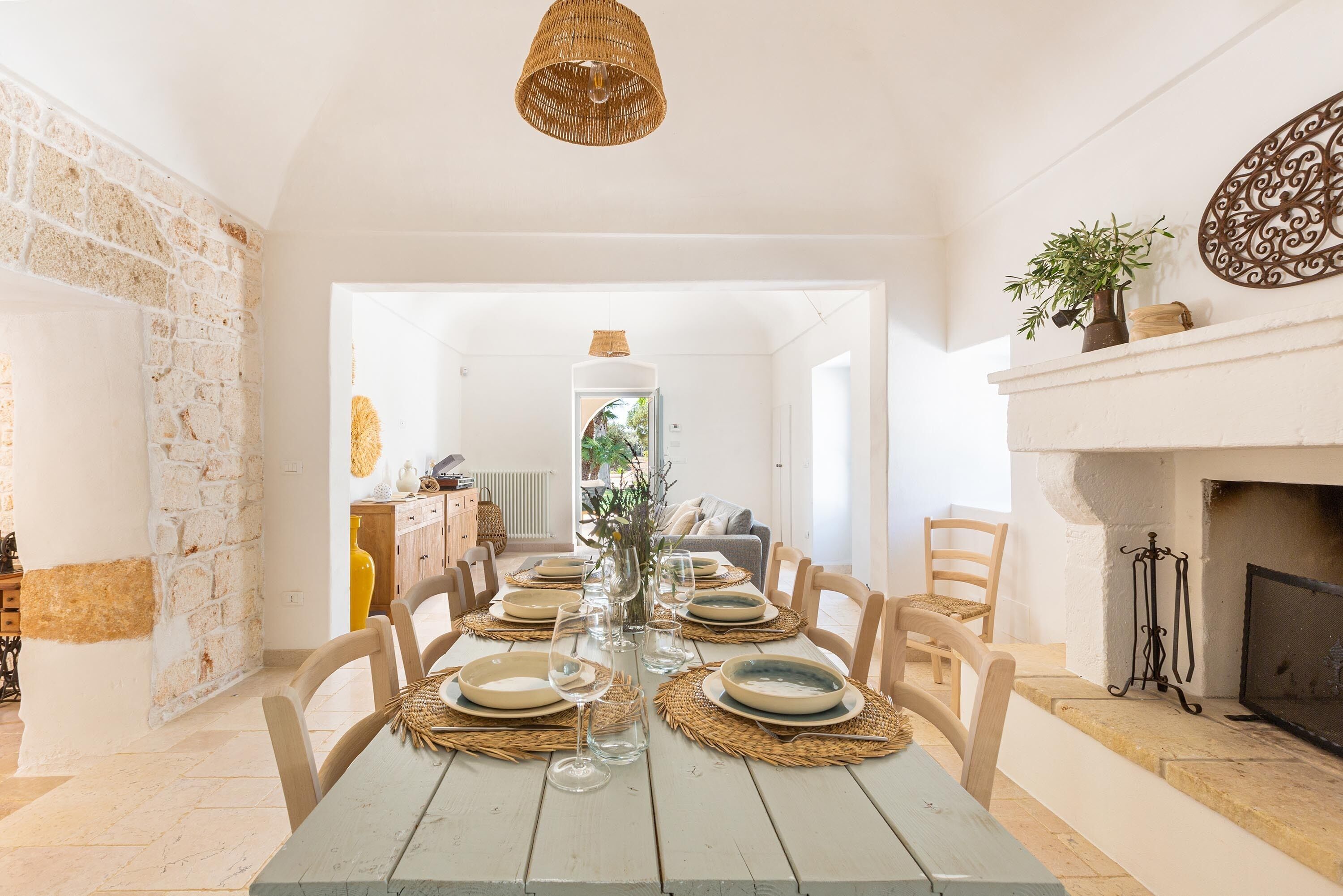 A dining area with a long table set for six, surrounded by chairs, with a fireplace and a decorative wall piece in the background.