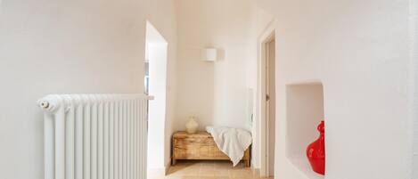 Un pasillo con un radiador blanco, un banco de madera con tela blanca y un jarrón rojo en la pared.