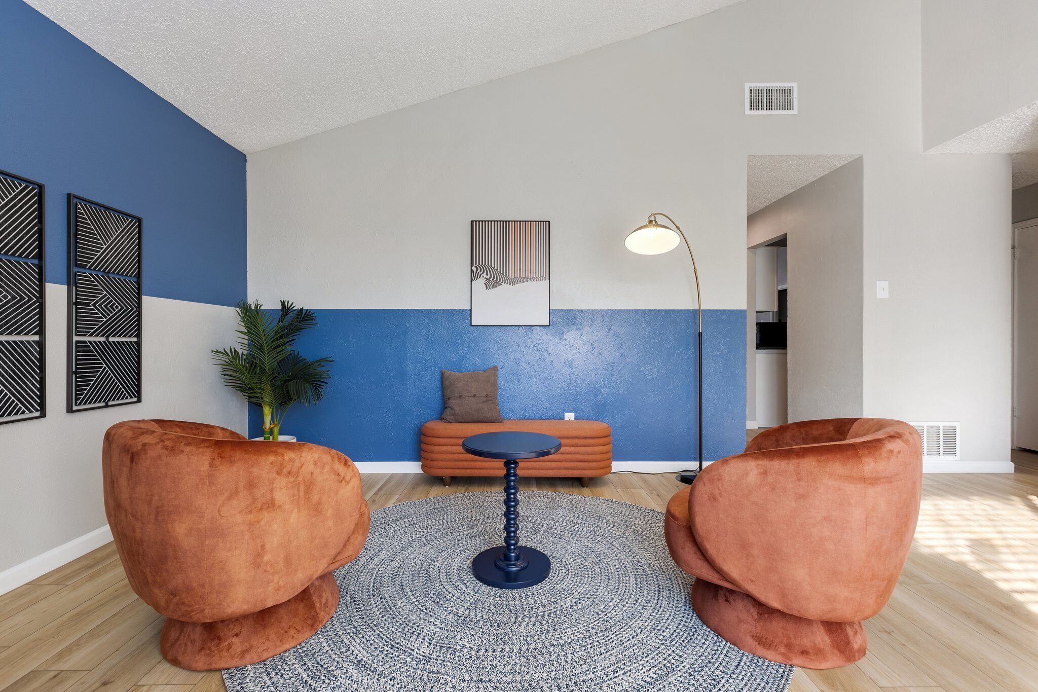 Een moderne woonkamer met een blauw-wit kleurenschema, met een ronde tafel, twee oranje fauteuils, een potplant en een aan de muur hangend kunstwerk.