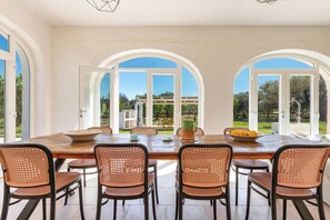 Comedor con una gran mesa de madera y sillas de mimbre, ventanas arqueadas amplias y vistas al jardín.