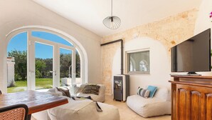 A spacious living area with a large window, a wooden table, and a television on a wooden cabinet. - 3483 Casale Amenola by Perle di Puglia (Ostuni)
