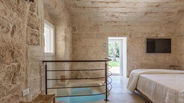 A stone-walled bedroom with a bed, a bench, a metal rack, and a window. - 3483 Casale Amenola by Perle di Puglia (Ostuni)