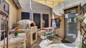 Une terrasse extérieure avec une cheminée en briques, une table avec des chaises, des guirlandes lumineuses et un parasol jaune.