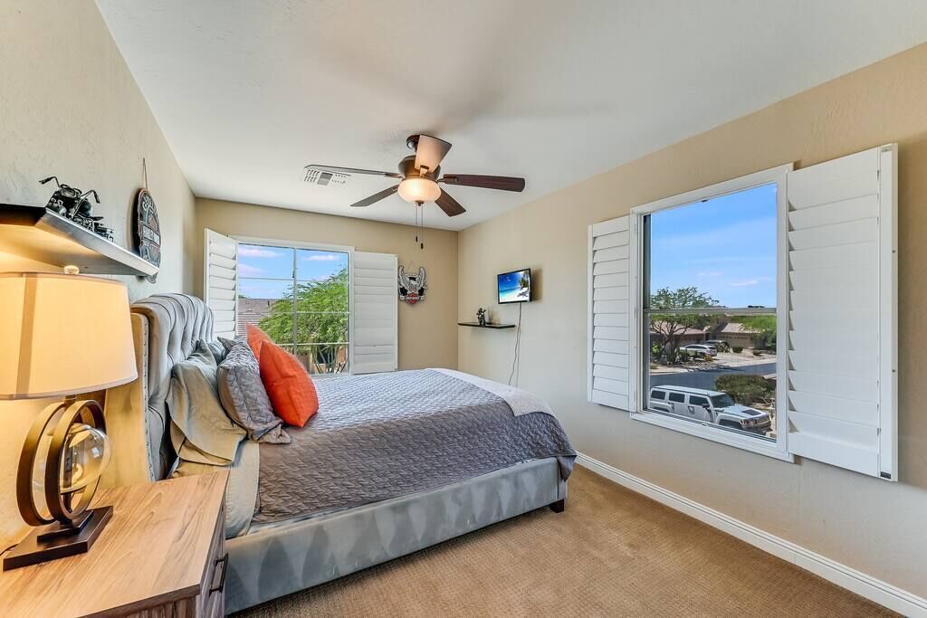 A bedroom with a bed, a ceiling fan, a TV, and a large window with shutters.