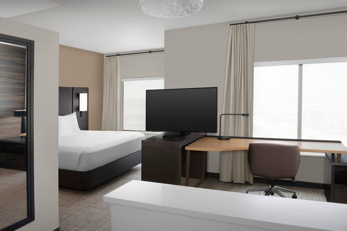 Ein Hotelzimmer mit Bett, Schreibtisch, Stuhl, Fernseher und großen Fenstern.