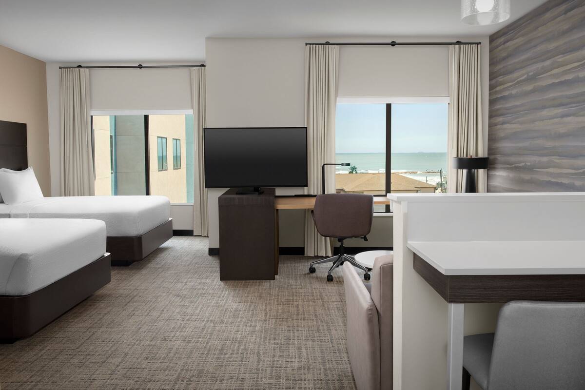 Ein Hotelzimmer mit zwei Betten, einem Schreibtisch, einem Stuhl, einem Fernseher und Blick auf den Strand.