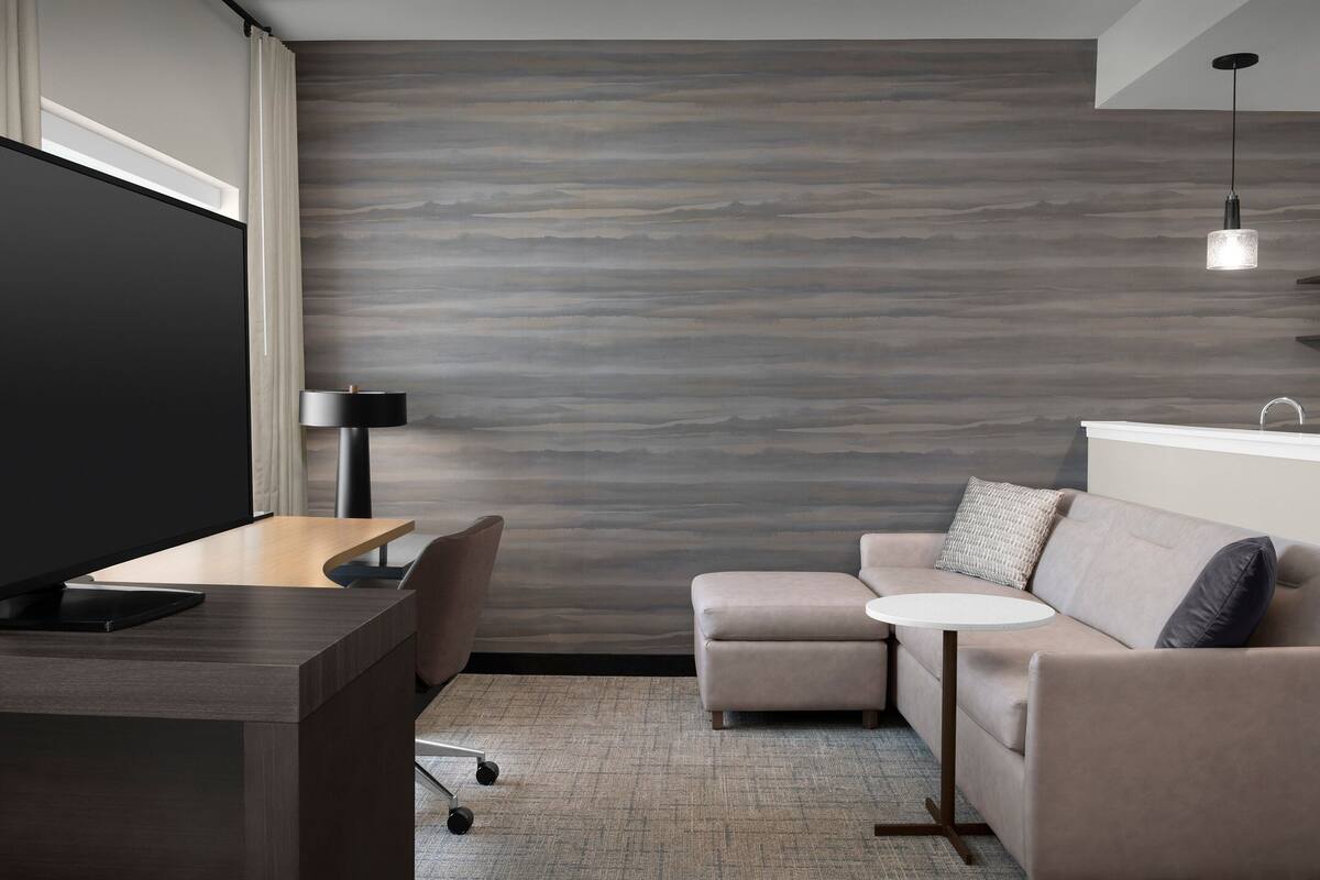 Ein modernes Hotelzimmer mit Flachbildfernseher, Schreibtisch, Sofa, rundem Tisch und Lampe.