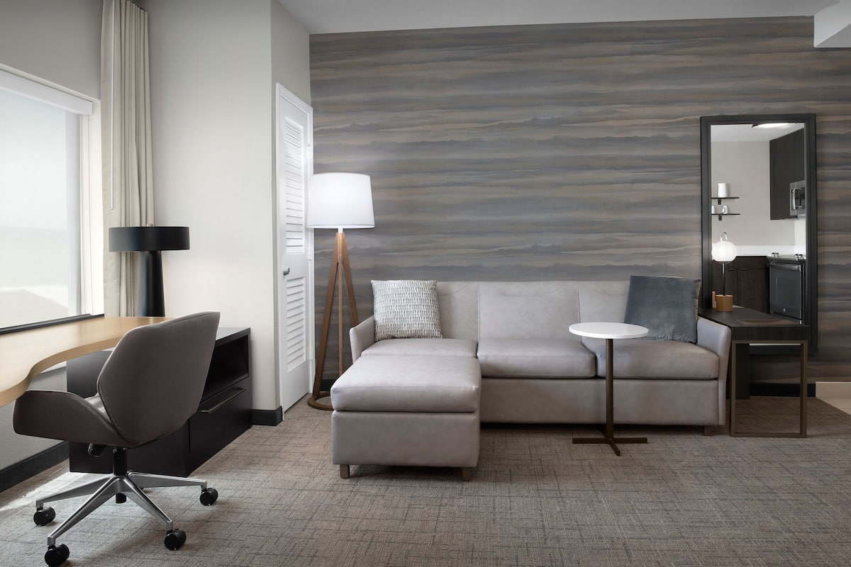 Ein modernes Hotelzimmer mit einer Eckcouch, einem Schreibtisch mit Stuhl, einem kleinen runden Tisch, einer Lampe und einem großen Spiegel.