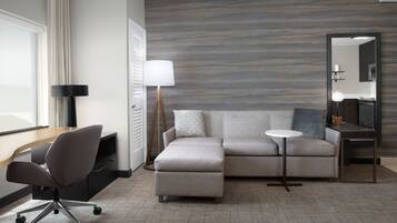 A modern hotel room with a sectional sofa, a desk with a chair, a small round table, a lamp, and a large mirror.