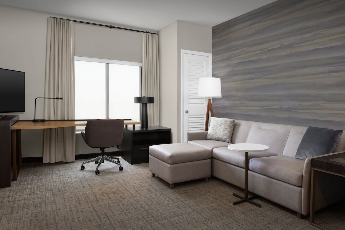Ein Hotelzimmer mit Schreibtisch, Bürostuhl, einer Couch und einem großen Fenster mit Vorhängen.