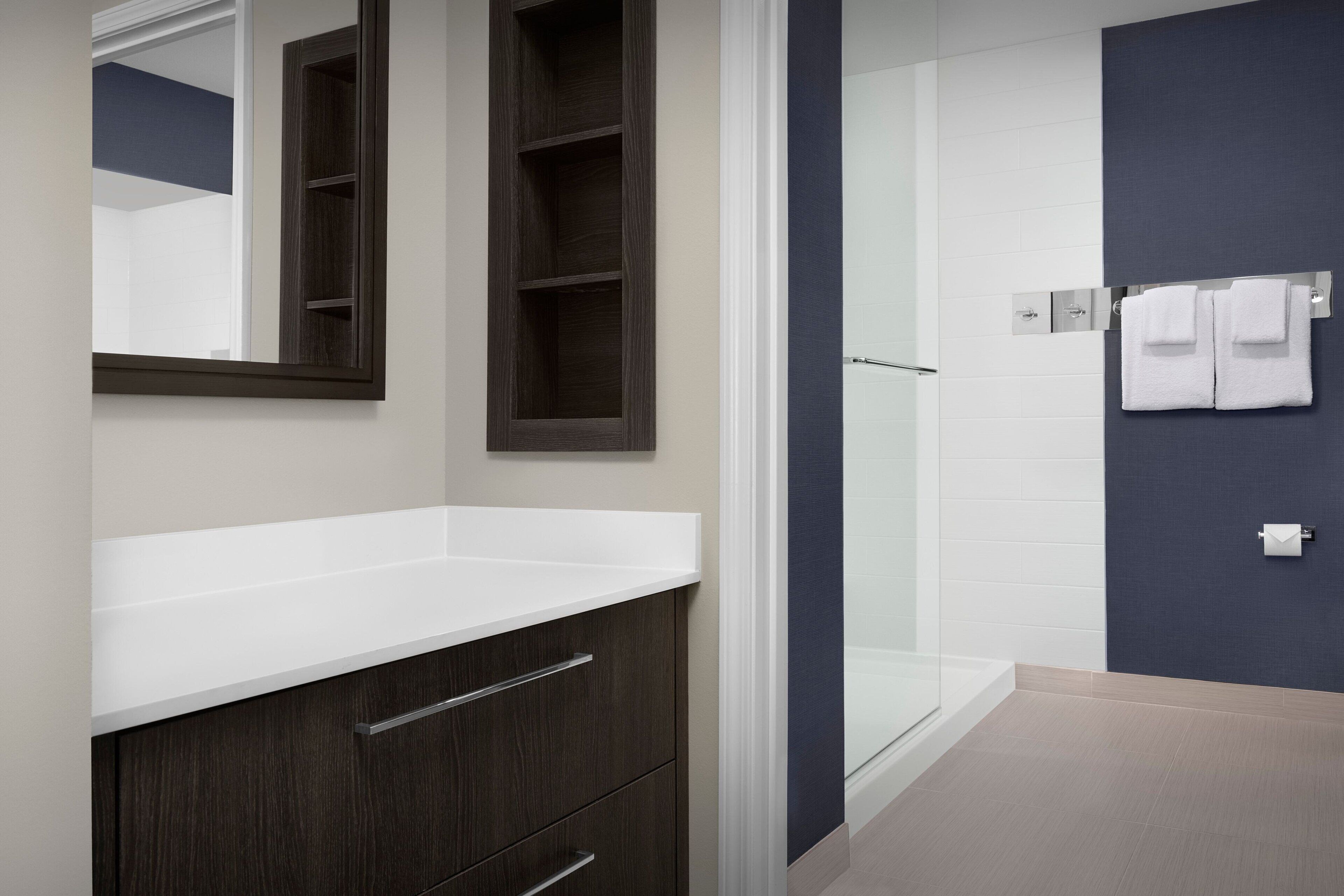 A modern bathroom with a dark wood vanity, a white countertop, a glass shower enclosure, and a dark blue accent wall.