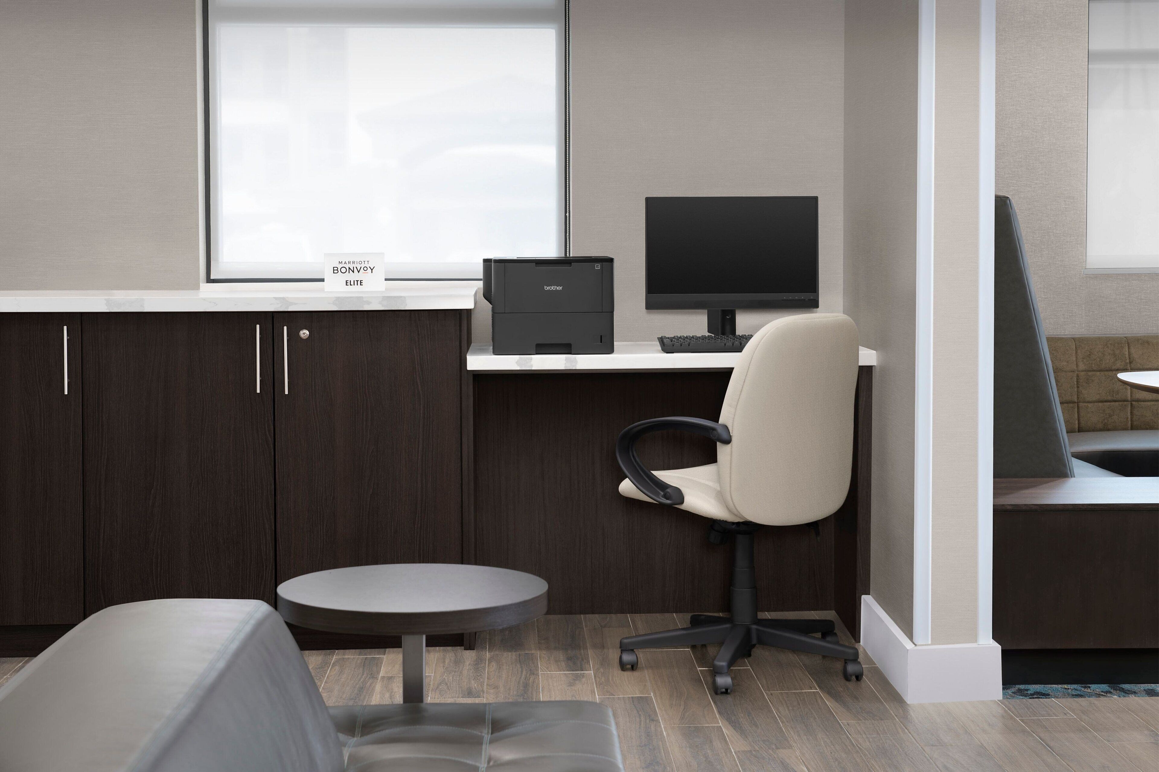 Et moderne kontor med et skrivebord, en stol og en computer. Der er et vindue med et skilt, der står "Marriott Bonvoy Elite".
