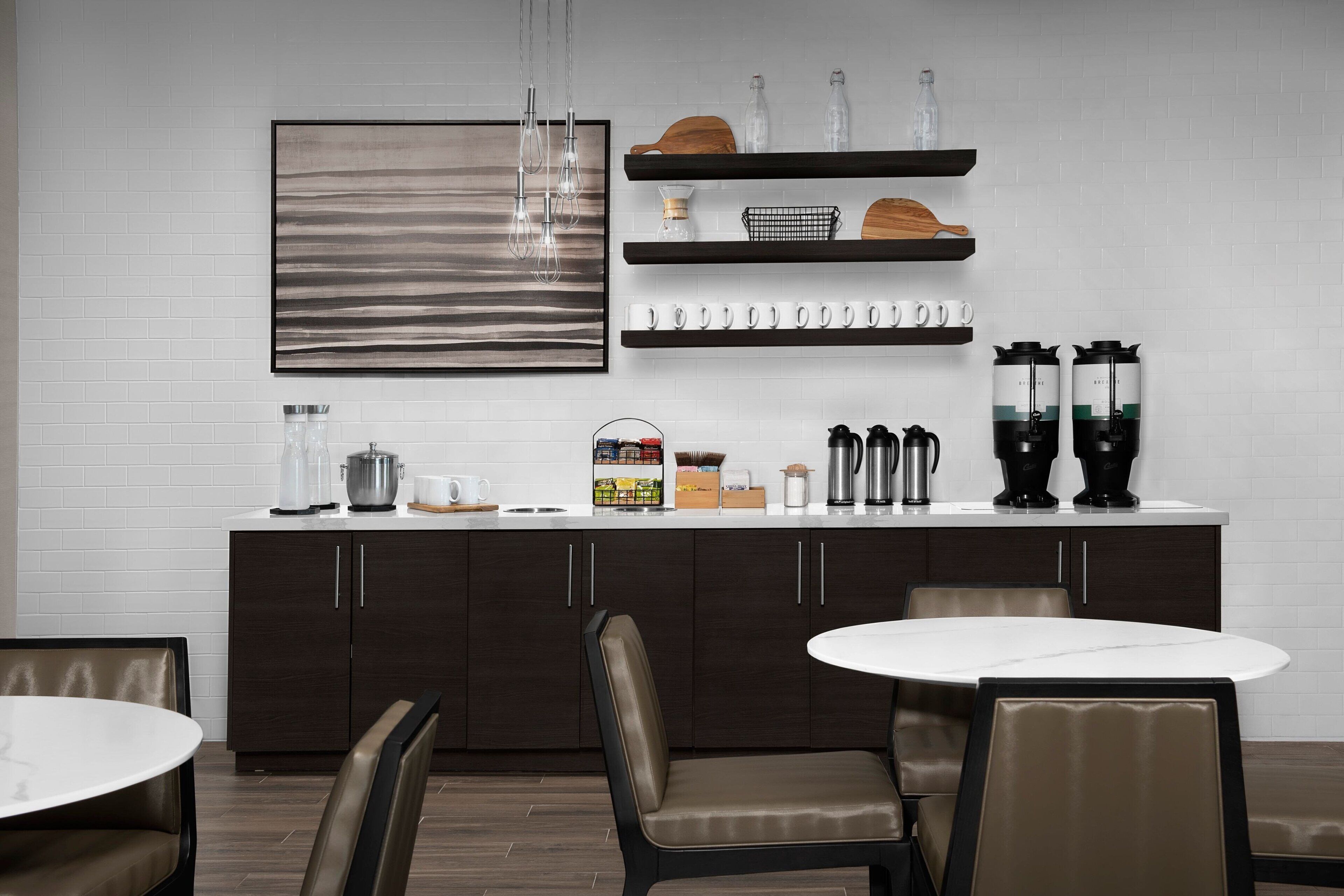 Eine moderne Küche mit weißer Ziegelrückwand, dunklen Holzschränken und einer weißen Arbeitsplatte. Auf der Arbeitsplatte befinden sich zwei Kaffeemaschinen, ein Wasserkocher und eine Kaffeemühle. Die Küche verfügt über einen runden weißen Tisch und zwei Stühle.