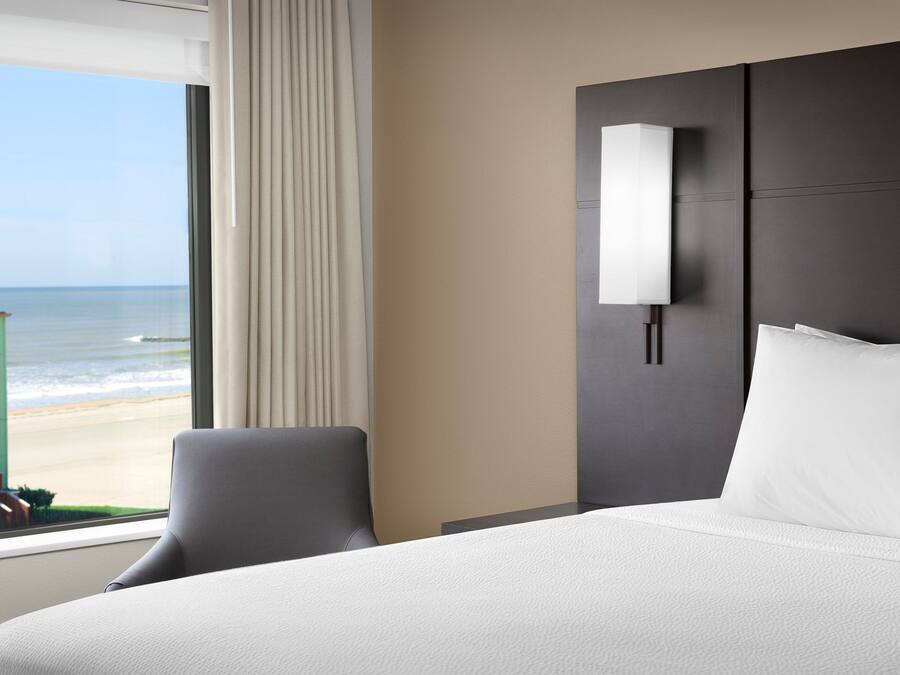 Ein Hotelzimmer mit einem großen Bett, einem Sessel, einem Nachttisch, einer Lampe und Blick auf den Strand.