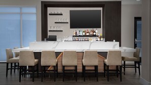 Un espace bar moderne doté d’un long comptoir, de nombreuses chaises, d’un grand téléviseur à écran plat, ainsi que de diverses bouteilles et verres exposés sur des étagères.