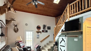 A cozy living room with a brown leather couch, a ceiling fan, wooden beams, and a staircase leading to an upper level.
