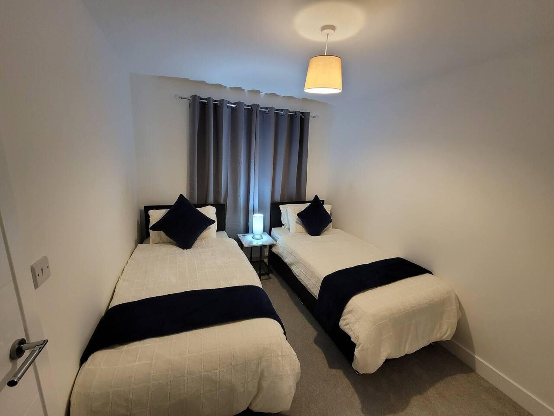 Una habitación de hotel con dos camas individuales, una mesita de noche con lámpara, una ventana con cortinas y una luz en el techo.