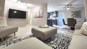 A modern living room with a flat-screen TV, a dining area with a table and chairs, and a kitchen in the background. - 6 guests, 4 min Walk to Private Beach, casino, bowling alley and much more ! 
 (Dorado)