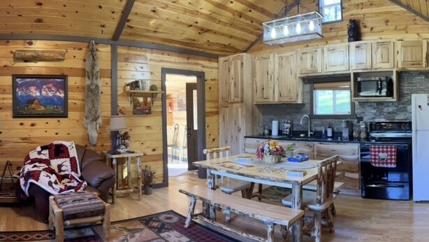 Ein gemütliches Blockhaus-Interieur mit einem Holzespeisetisch, Bänken, einem Kamin und einer Küchenzeile mit Mikrowelle und Herd.