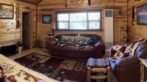 Ein gemütliches Blockhäuschen-Interieur mit Holzbalkendecke, großem Fenster, braunem Ledersofa, Holzstuhl und Teppich mit Tiermotiv.