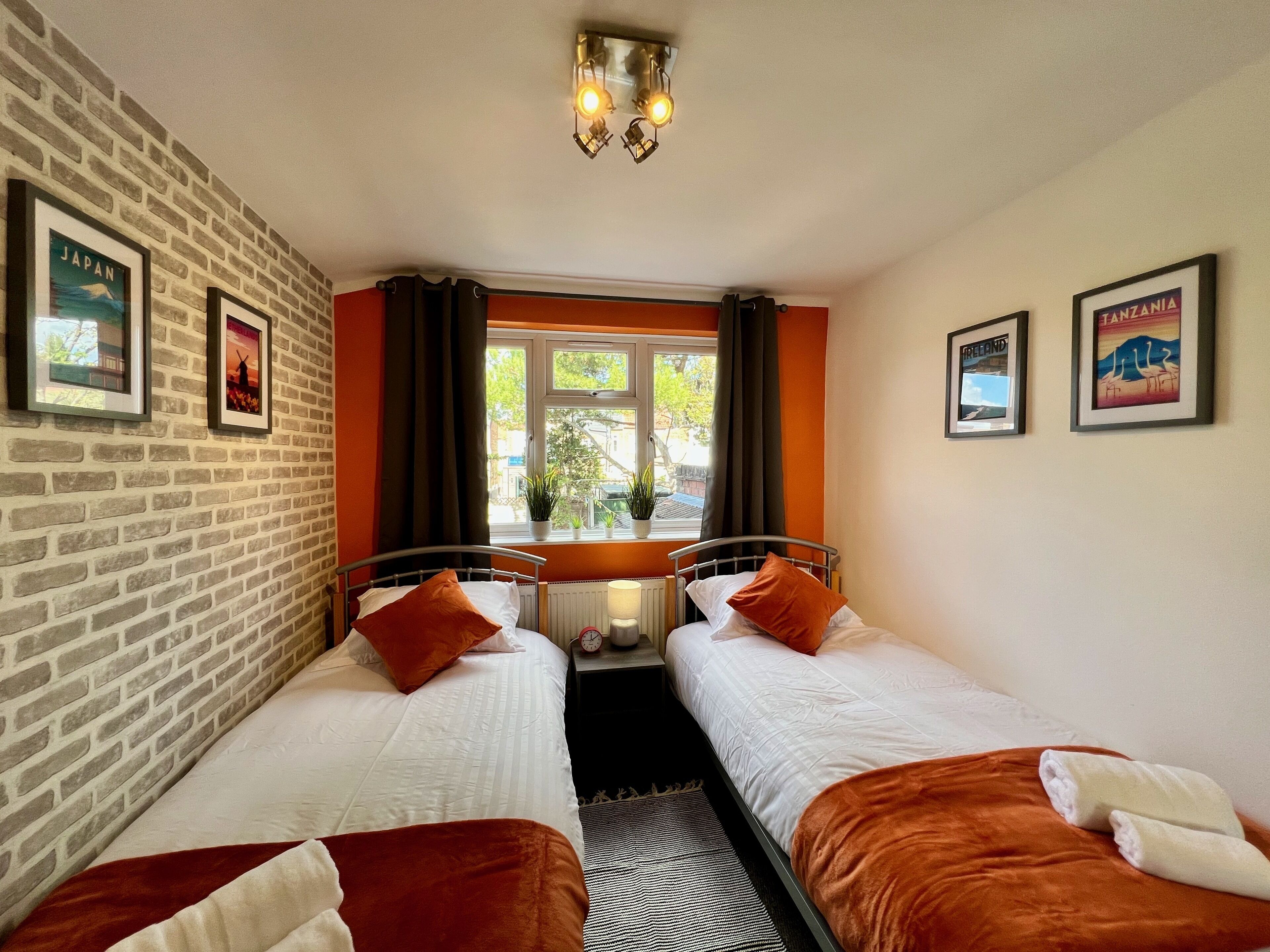 A room with two beds, a brick wall, framed pictures, a window with curtains, and a ceiling light fixture.