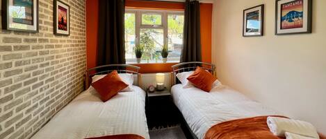 A room with two beds, a brick wall, framed pictures, a window with curtains, and a ceiling light fixture.