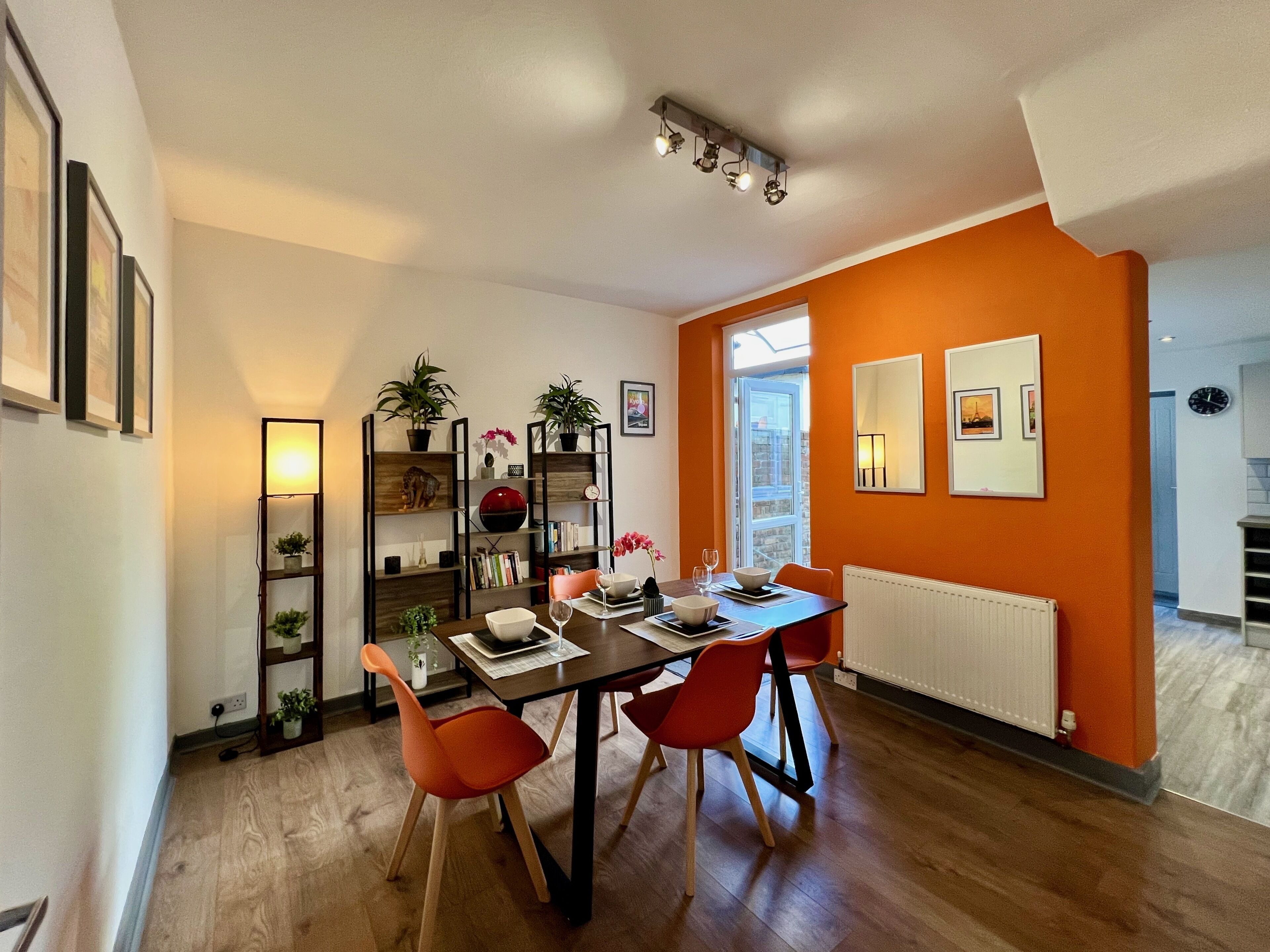 Un comedor con una mesa de madera, sillas naranjas y una estantería con plantas.