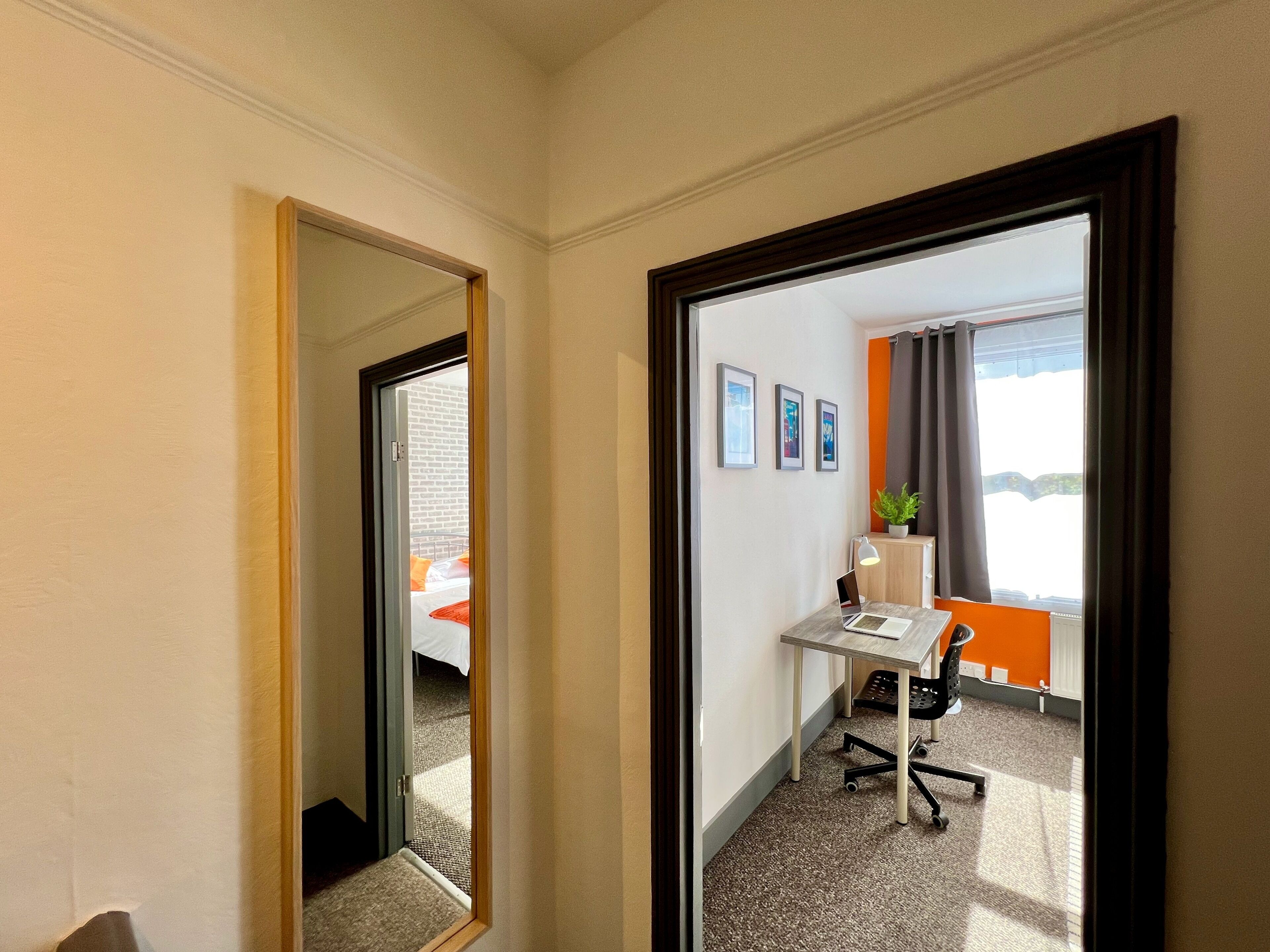 A hotel room with a desk, chair, and a bed with orange bedding. There are three framed pictures on the wall and a potted plant on the desk.