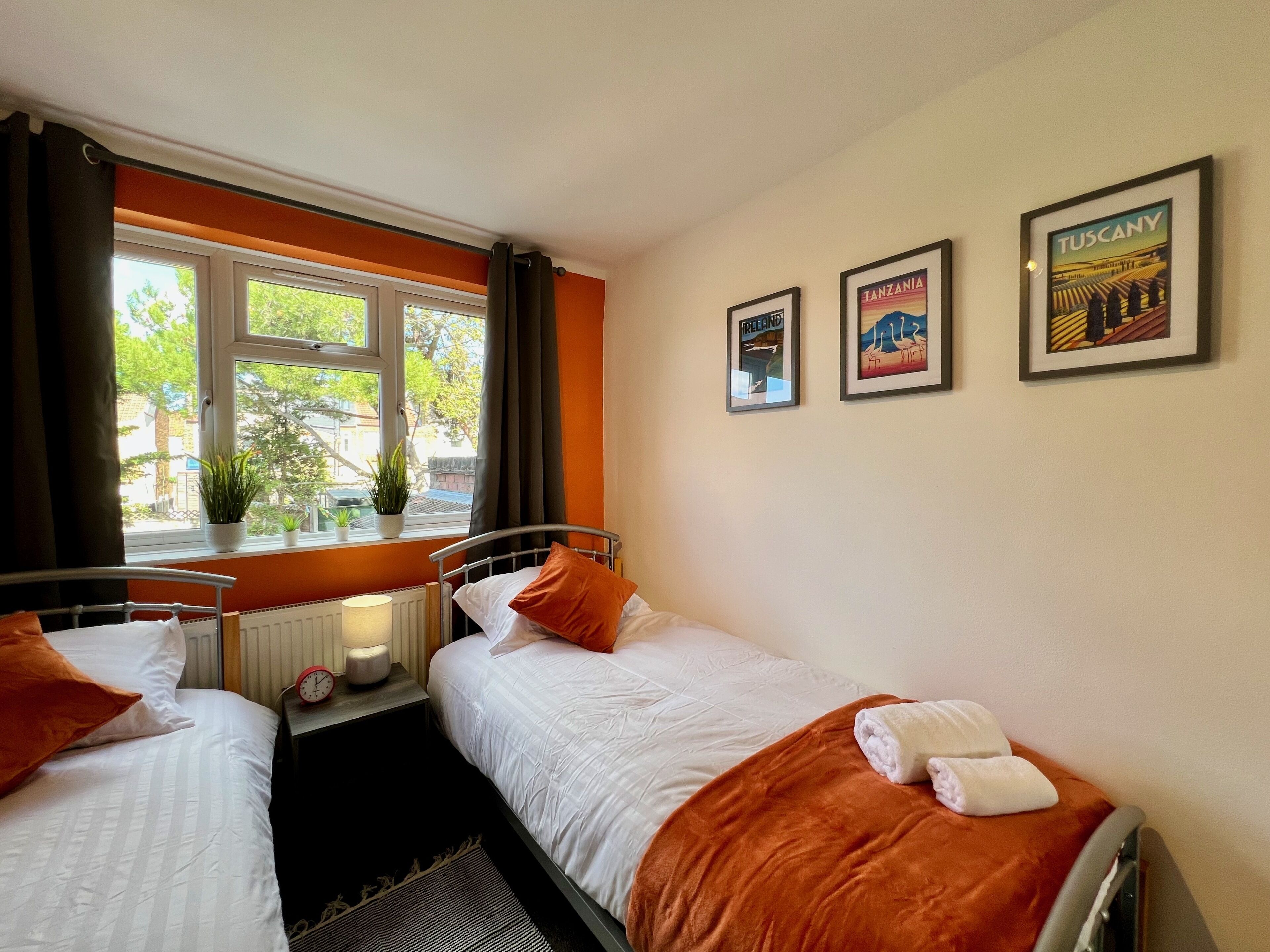 Camera d'albergo con due letti singoli, una finestra con tende, un termosifone, un comodino con una lampada e quadri appesi alle pareti.